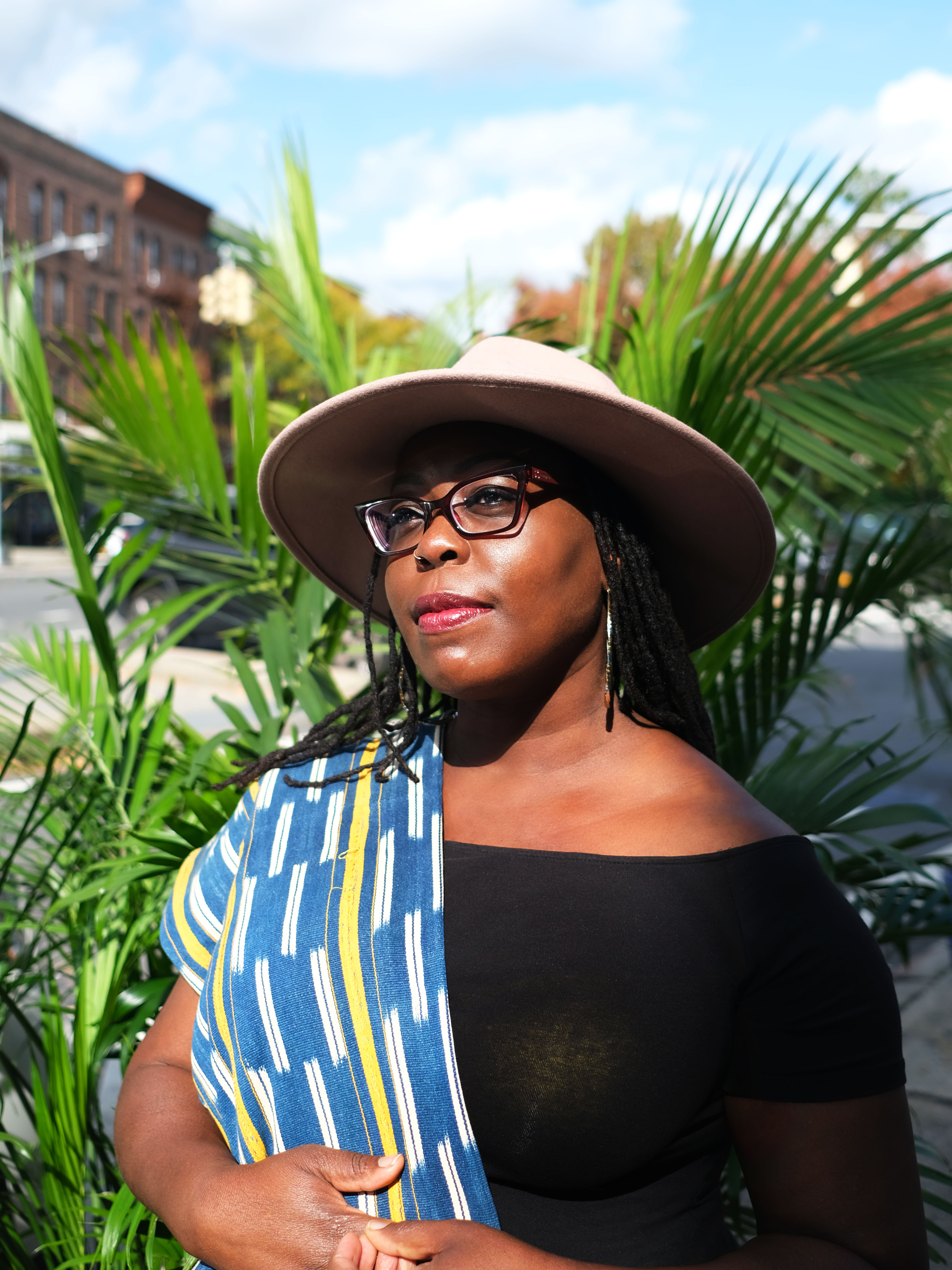 Portrait of Laylah Amatullah Barrayn from torso up wearing a hat looking toward left side of frame in front of palm fronds