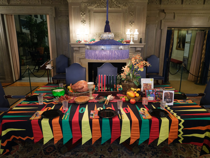 A Kwanzaa dinner table setting at the George Eastman Museum featuring a cooked turkey, a kinara with lit candles, fruit, and books, all illuminated by a purple fringed lampshade.