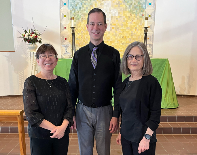 Three adults stand indoors in front of an altar with green cloth and candles at the George Eastman Museum. Two women in black tops flank a man in a black vest, shirt, and tie. They are all smiling at the camera, perhaps ready to enjoy Keyboard Duets of Depth and Diversion.