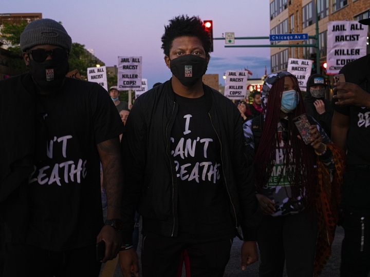 Two people at a march wearing t-shirts with the words I Can't Breathe, among a crowd of other people holding several signs with the words "Jail All Racist Killer Cops" 