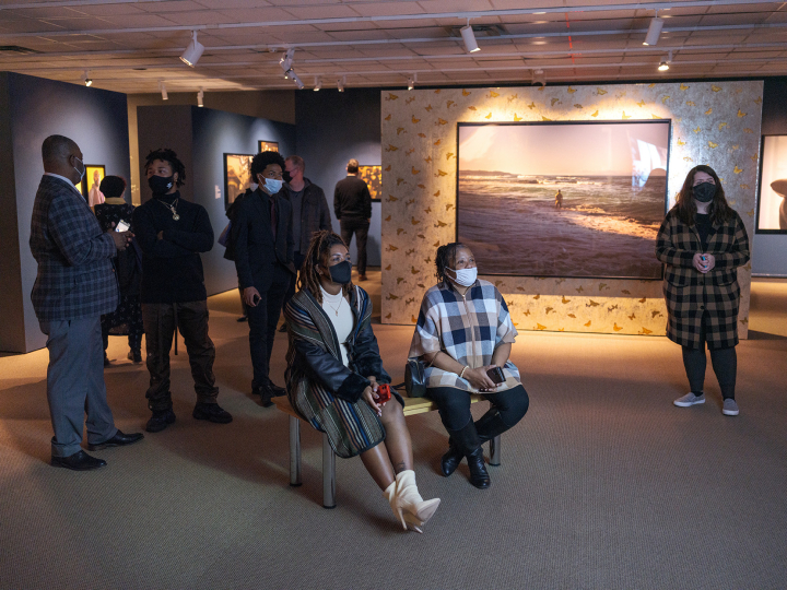 Exhibition gallery featuring large color photographs with four adults, two seated and one standing, looking at something off the right side of the frame