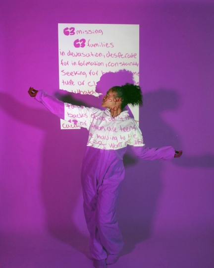 Young teenage girl of color in front of a poem projected on a film. Says "63 missing and 63 families" and they are dressed in purple