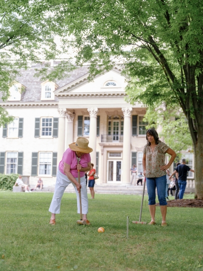 Two adults playing croquet on the front lawn of the George Eastman Museum. The museum's historic mansion is visible in the background.