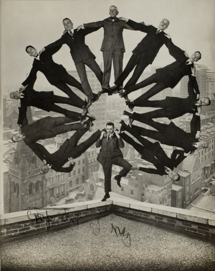 Photo of a man posed on the corner of a rooftop with an arc of eleven suited men balanced on his shoulders