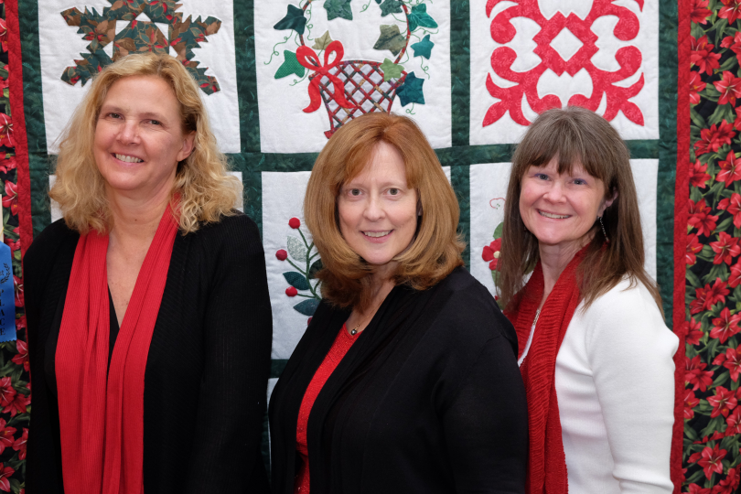 Women's vocal jazz music trio LumenEssence. Three women stand in front of a festive quilt with red and green patterns. They are smiling and each wears a red scarf, black or white tops, and have shoulder-length hair.