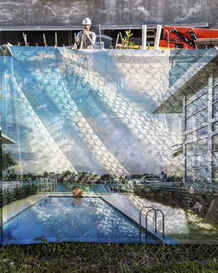 Construction cloth printed with an image of a swimming pool, covering chainlink fence, with construction worker visible at top of frame behind fence