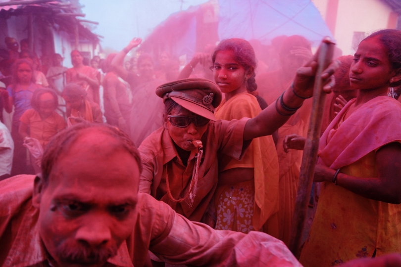 Crowd of people outdoors with one central figure blowing a whistle and another young woman looking into the camera, with magenta powder (gulal?) covering everything in view