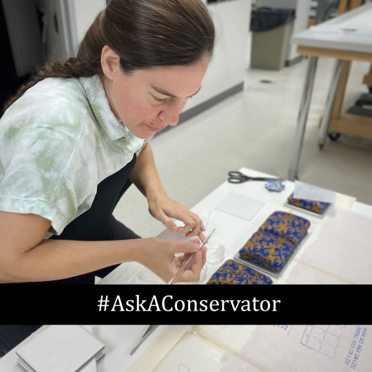 A conservator uses a thin tool to work on a small, patterned blue and gold decorative object on a studio table, captured for Ask a Conservator Day at the George Eastman Museum, with the text #AskAConservator overlaid at the bottom.