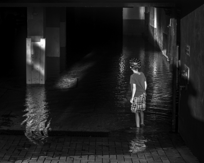 Dark interior of flooded parking garage with child seen from behind standing in water wearing a bicycle helmet