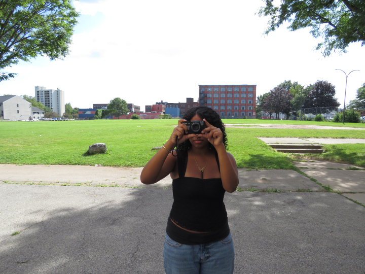 A person in a black top and blue jeans stands outdoors in a park, holding a camera up to their face as they take a photo. Green grass, trees, and buildings are visible in the background—capturing the curiosity and creative spirit celebrated by Click & Tell, a summer photography club for teens that fosters storytelling through images. This scene reflects the vibrant energy found in the Gallery Obscura exhibition From Where I Stand: Click & Tell: Photography Club at the Trenton and Pamela Jackson R-Center, wh