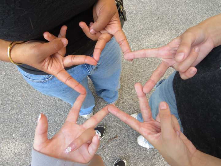 Four people form a star shape by touching their fingers together while standing in a circle on a paved surface—a perfect moment of connection and creativity captured by Click & Tell, a summer photography club for teens that celebrates curiosity and imagination as we look ahead to 2027.