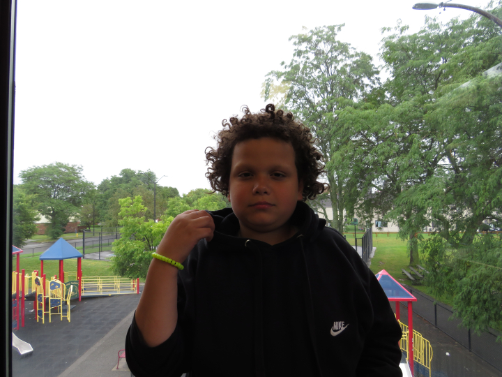 A child with curly hair wearing a black Nike hoodie stands indoors in front of a window, with a playground and trees visible outside on a cloudy day—a quiet moment that reflects curiosity and the power of storytelling through images. This photograph is part of the Gallery Obscura exhibition From Where I Stand: Click & Tell: Photography Club at the Trenton and Pamela Jackson R-Center, which features a vibrant collection of photographs by participating students that showcase the resilience and imagination of 