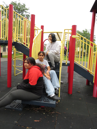 Three young people sit and one kneels on a playground structure with red and yellow railings. Trees and grass are visible in the background, adding to the relaxed and casual scene. This moment, reminiscent of the creative spirit fostered by Click & Tell—a summer photography club for teens that celebrates creativity—shows friends enjoying time together and capturing memories.