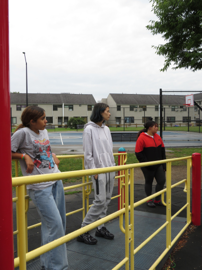 Three people stand on a playground structure near a basketball court and fence, with apartment buildings in the background. The sky is overcast, and the group looks in different directions. Part of the Gallery Obscura exhibition “From Where I Stand: Click & Tell: Photography Club at the Trenton and Pamela Jackson R-Center,” this scene highlights Click & Tell—a summer photography club for teens that celebrates creativity, curiosity, and the power of storytelling through images—showcasing the resilience and i