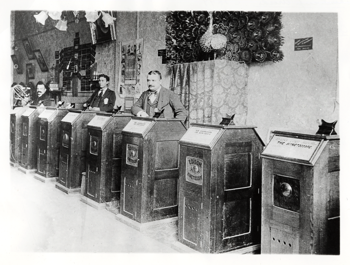 Three men among a line of eight Edison Kinetoscopes