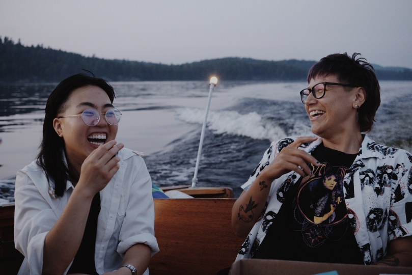 Two people laugh joyfully while sitting on a boat during a calm evening ride, with water and a forested shoreline in the background.