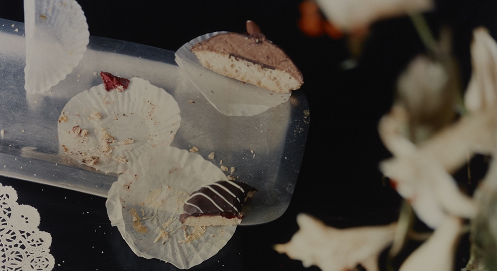Photograph by Laura Letinsky depicting partially eaten pastries and crumbs on paper doilies on a dark table, with blurry flowers in right foreground