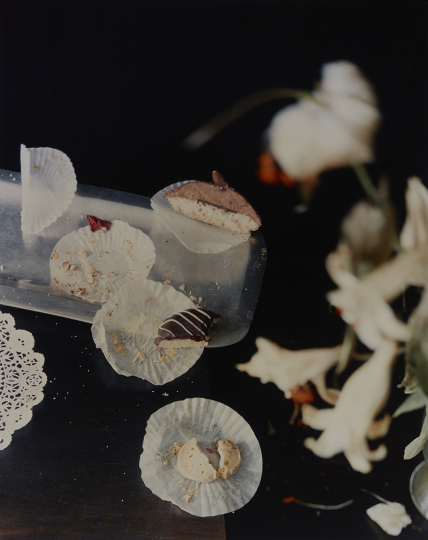 Photograph by Laura Letinsky depicting partially eaten pastries and crumbs on paper doilies on a dark table, with blurry flowers in right foreground