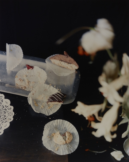 Photograph by Laura Letinsky depicting partially eaten pastries and crumbs on paper doilies on a dark table, with blurry flowers in right foreground