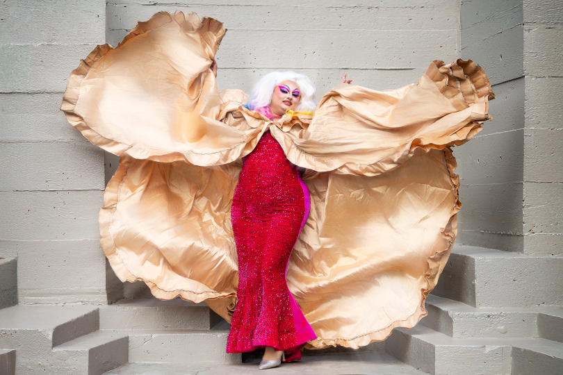 A drag performer poses confidently on concrete steps wearing a sparkling red gown and a dramatic, flowing gold cape with voluminous ruffles.