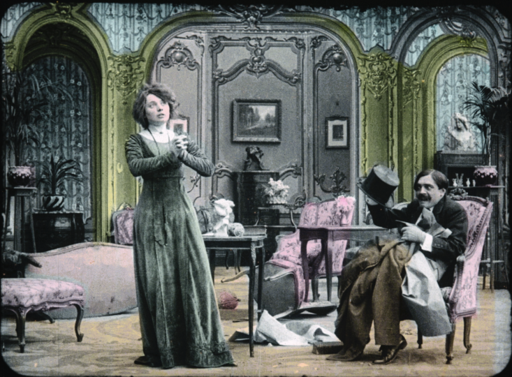 Hand painted film still that was originally black and white. A 19th c woman has her hands clasped while a gentleman sitting down in the parlor looks at her. 