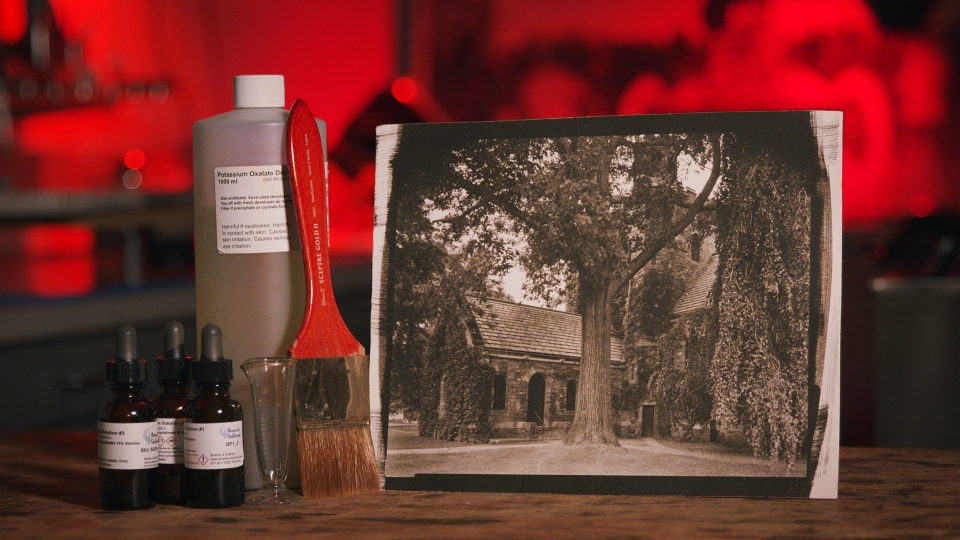 Display of photography materials which includes potassium 1000 ml bottle, a large painting brush, a small measuring vessel, and three contains of solution. Next to the materials is a vintage photo of a large tree in front of an old stone church-like structure. In the background is a blurred image of a photographic darkroom.  