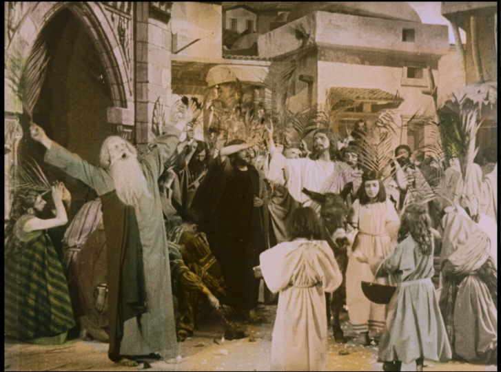 A hand painted film still that was originally black and white. An old man holding out a palm exclaims as Jesus Christ on a donkey passes him.  