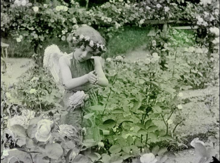 A hand painted film still that was black and white. A little girl poses as cupid in a rose garden.