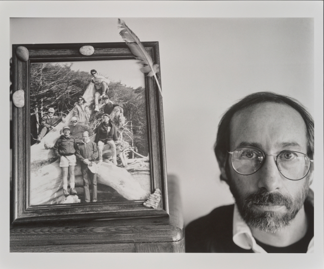 A black-and-white portrait of a man with glasses by Albert J. Winn, positioned next to a framed photograph of a group of people posing on a large fallen tree from his 1991 series, My Life Until Now.