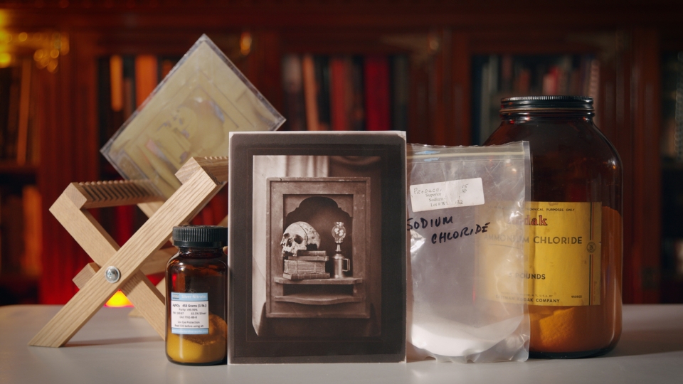 Display of photographic materials including wooden drying rack, bottle of silver nitrate, bag of sodium chloride, bottle of ammonium chloride, and salt print photograph of a skull and books