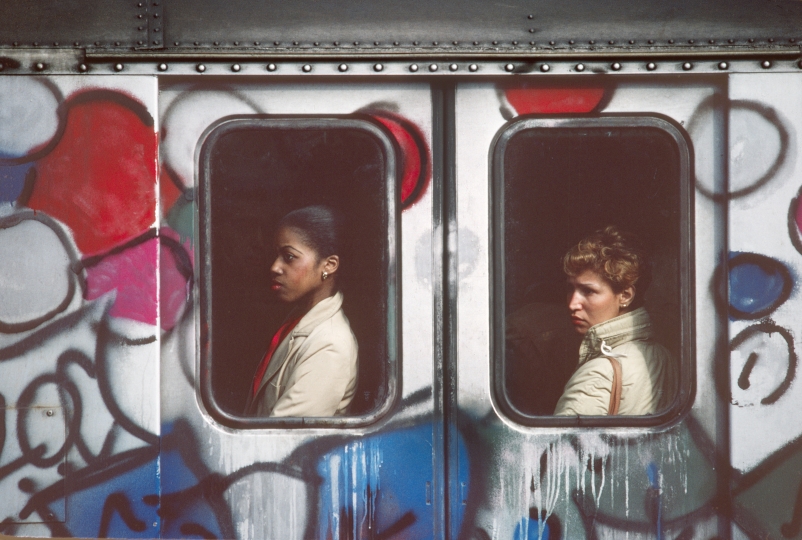 Graffiti-covered subway car from the outside, with a person visible in each of the two windows