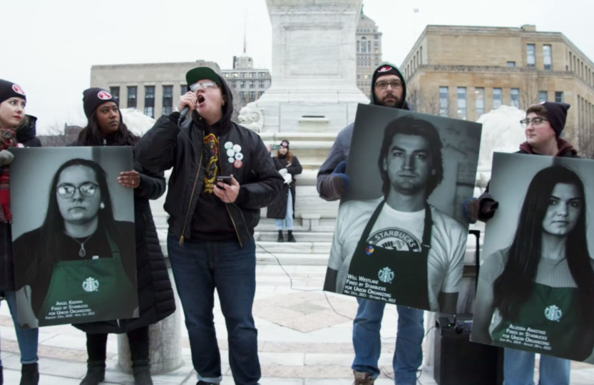 	 A group of people stand outside holding large posters with black-and-white photos of individuals wearing Starbucks aprons, while one person speaks into a microphone. This powerful scene is featured in Baristas vs. Billionaires (DCP), directed by the Labor Film Series at the Dryden Theater, US 2025, 79 min.