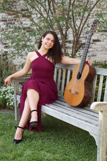 Maya Loncar, guitarist, in a long dress and sitting on a bench with her guitar outside