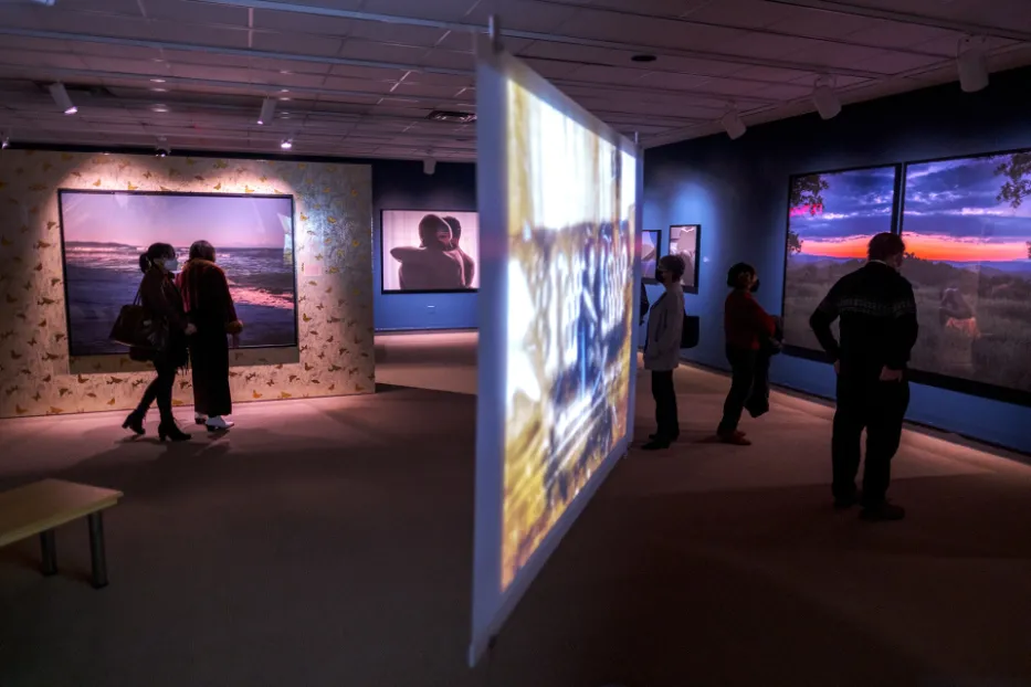 Exhibition gallery featuring large color photographs and a projection screen with five adults looking at art throughout the space