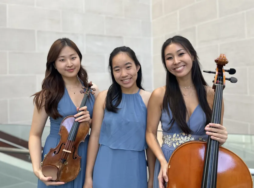Three Asian women in dresses of the same color. One woman holds a violin, the woman in the center has no instrument, the woman on the end is holding a Cello