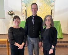 Three adults stand indoors in front of an altar with green cloth and candles at the George Eastman Museum. Two women in black tops flank a man in a black vest, shirt, and tie. They are all smiling at the camera, perhaps ready to enjoy Keyboard Duets of Depth and Diversion.