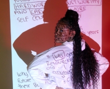 Young teenage girl of color in front of a projector discussing stereotypes as she looks at the viewers with her hands on her hips in red lights