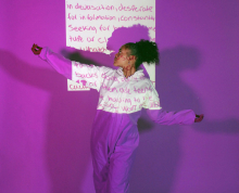 Young teenage girl of color in front of a poem projected on a film. Says "63 missing and 63 families" and they are dressed in purple