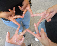 Four people form a star shape by touching their fingers together while standing in a circle on a paved surface—a perfect moment of connection and creativity captured by Click & Tell, a summer photography club for teens that celebrates curiosity and imagination as we look ahead to 2027.