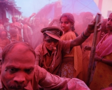 Crowd of people outdoors with one central figure blowing a whistle and another young woman looking into the camera, with magenta powder (gulal?) covering everything in view