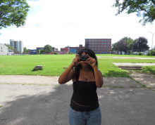 A person in a black top and blue jeans stands outdoors in a park, holding a camera up to their face as they take a photo. Green grass, trees, and buildings are visible in the background—capturing the curiosity and creative spirit celebrated by Click & Tell, a summer photography club for teens that fosters storytelling through images. This scene reflects the vibrant energy found in the Gallery Obscura exhibition From Where I Stand: Click & Tell: Photography Club at the Trenton and Pamela Jackson R-Center, wh