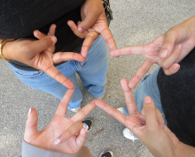 Four people form a star shape by touching their fingers together while standing in a circle on a paved surface—a perfect moment of connection and creativity captured by Click & Tell, a summer photography club for teens that celebrates curiosity and imagination as we look ahead to 2027.