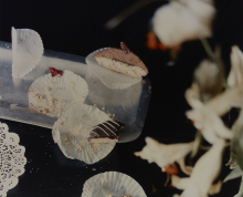 Photograph by Laura Letinsky depicting partially eaten pastries and crumbs on paper doilies on a dark table, with blurry flowers in right foreground