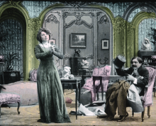 Hand painted film still that was originally black and white. A 19th c woman has her hands clasped while a gentleman sitting down in the parlor looks at her. 