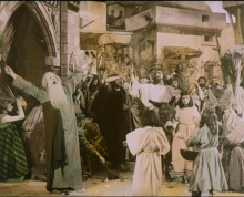 A hand painted film still that was originally black and white. An old man holding out a palm exclaims as Jesus Christ on a donkey passes him.  