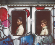 Graffiti-covered subway car from the outside, with a person visible in each of the two windows