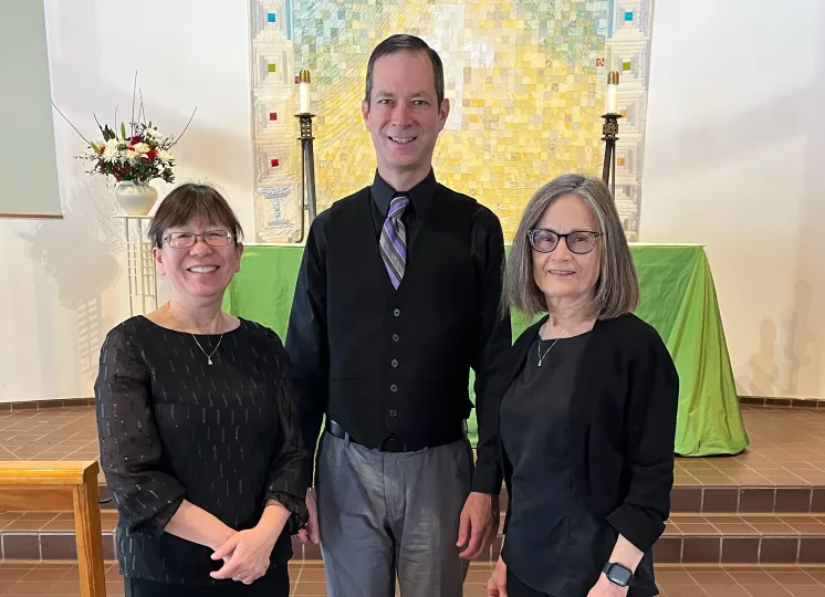 Three adults stand indoors in front of an altar with green cloth and candles at the George Eastman Museum. Two women in black tops flank a man in a black vest, shirt, and tie. They are all smiling at the camera, perhaps ready to enjoy Keyboard Duets of Depth and Diversion.