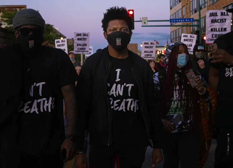 Two people at a march wearing t-shirts with the words I Can't Breathe, among a crowd of other people holding several signs with the words "Jail All Racist Killer Cops" 