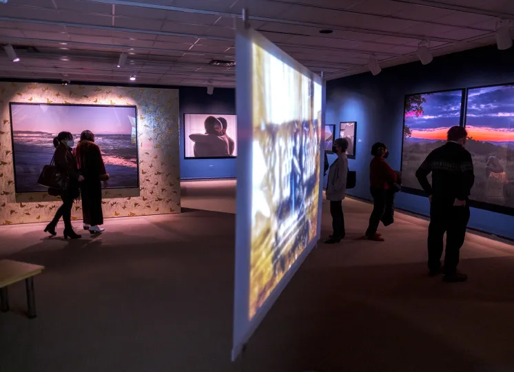 Exhibition gallery featuring large color photographs and a projection screen with five adults looking at art throughout the space