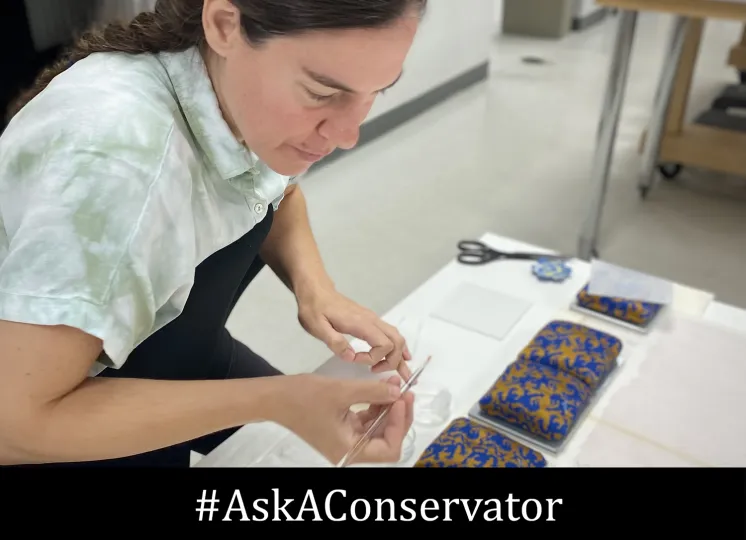 A conservator uses a thin tool to work on a small, patterned blue and gold decorative object on a studio table, captured for Ask a Conservator Day at the George Eastman Museum, with the text #AskAConservator overlaid at the bottom.